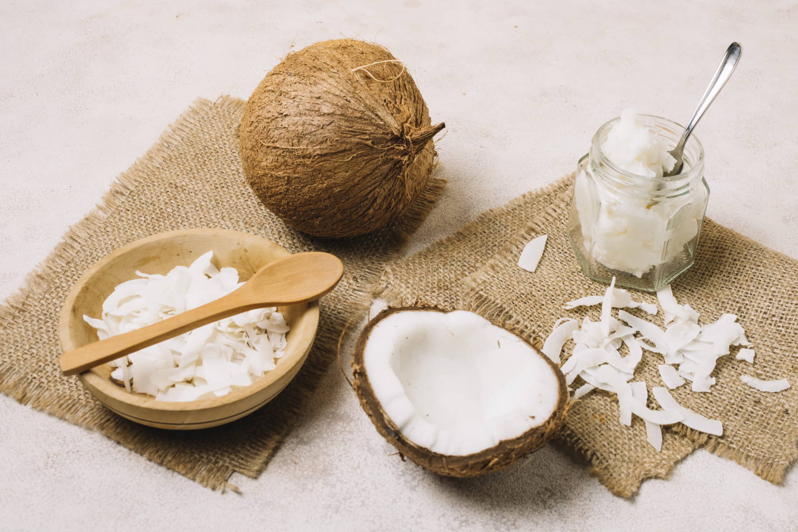 This image showcases various coconut products displayed on a sackcloth background. The setup includes a whole coconut, a halved coconut with the white flesh visible, a jar of coconut oil with a spoon, a bowl of shredded coconut, and scattered coconut flakes.