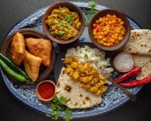 An assortment of delicious Indian vegetarian dishes served on a decorative tray, featuring samosas, chickpea curry, flatbread, spiced rice, and vibrant garnishes like fresh chili and herbs.